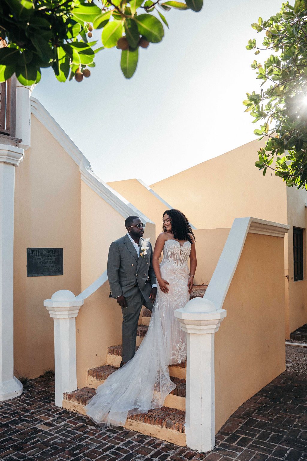 Kayla and Anthony stand together on the steps of a building at Avila Beach Hotel, surrounded by greenery in bright sunlight