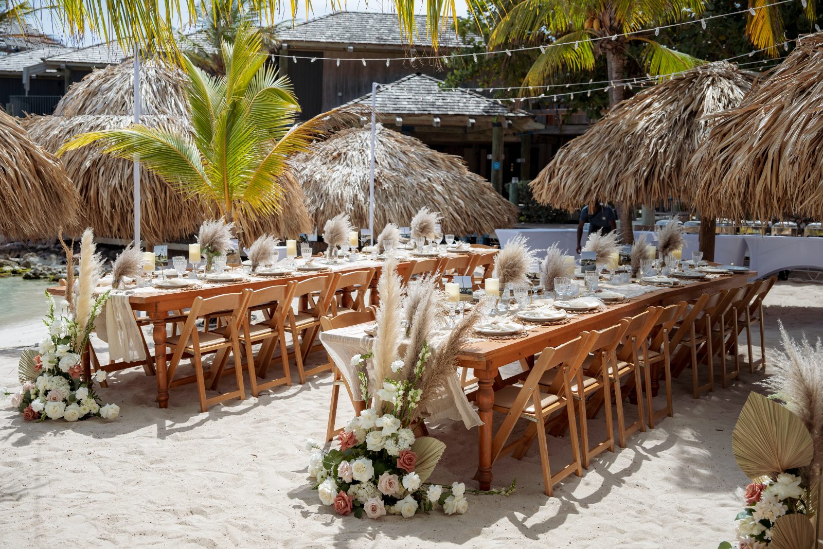 Kayla and Anthony's wedding reception is set on the beach at Avila Beach Hotel, featuring decorated tables under thatched umbrellas with tropical plants