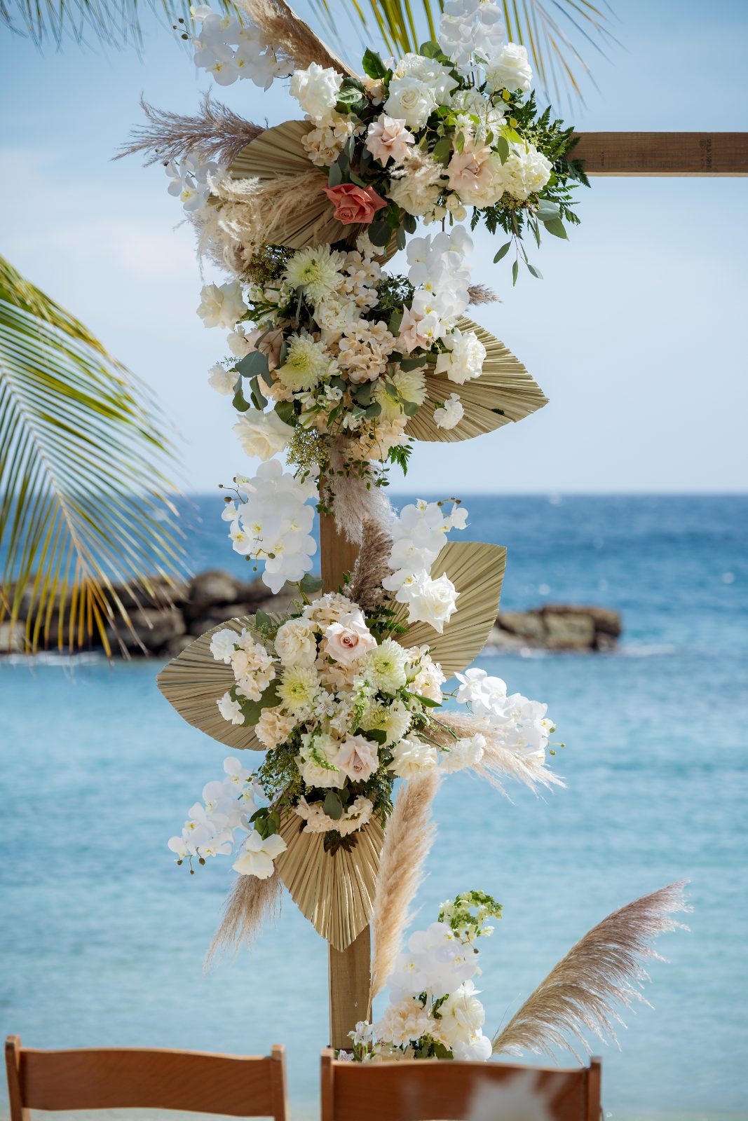 Kayla and Anthony stand in front of a floral arch with a beach backdrop at the Avila Beach Hotel during their wedding ceremony