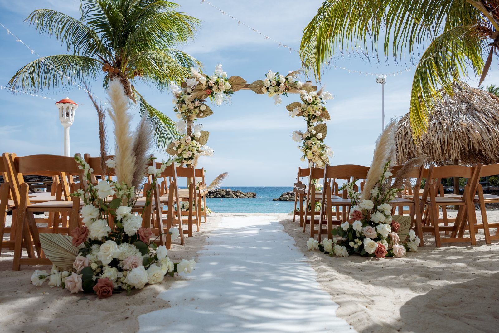 Kayla and Anthony's wedding ceremony takes place at Avila Beach Hotel, featuring an arch decorated with flowers and chairs set on the sandy beach