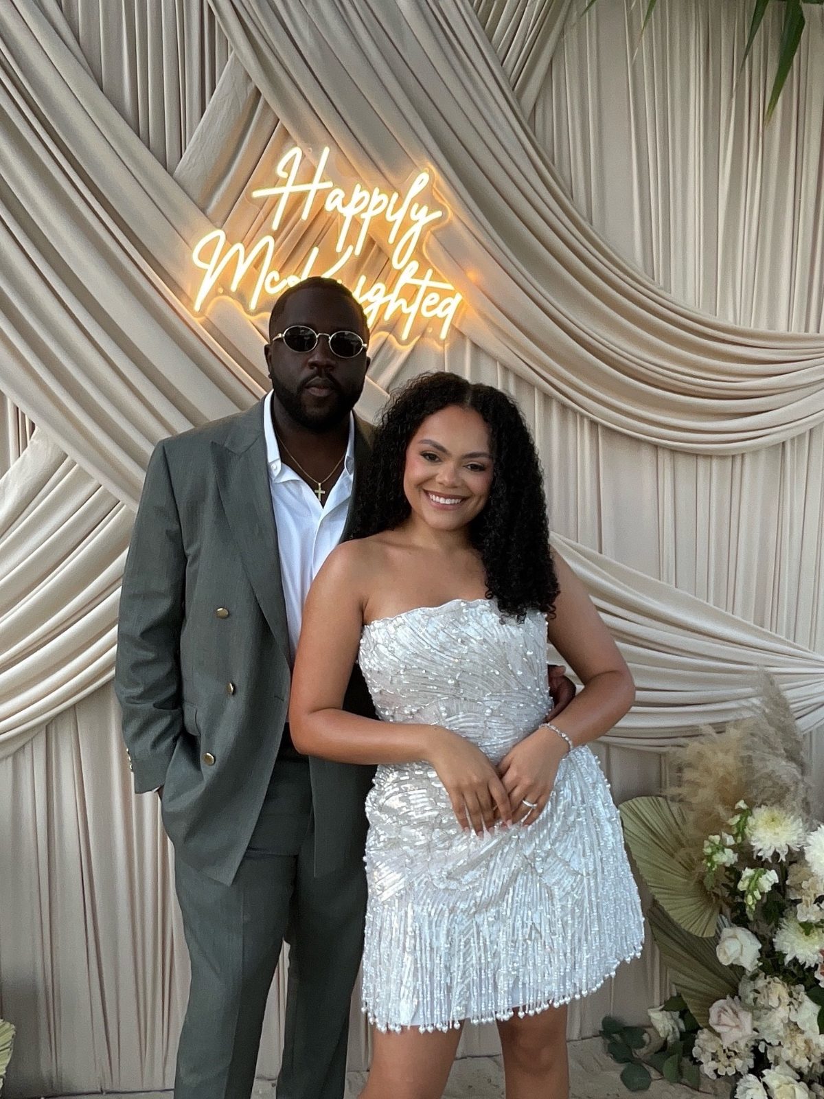 Kayla and Anthony stand together at their wedding reception at Avila Beach Hotel, with a "Happily Married" sign in the background