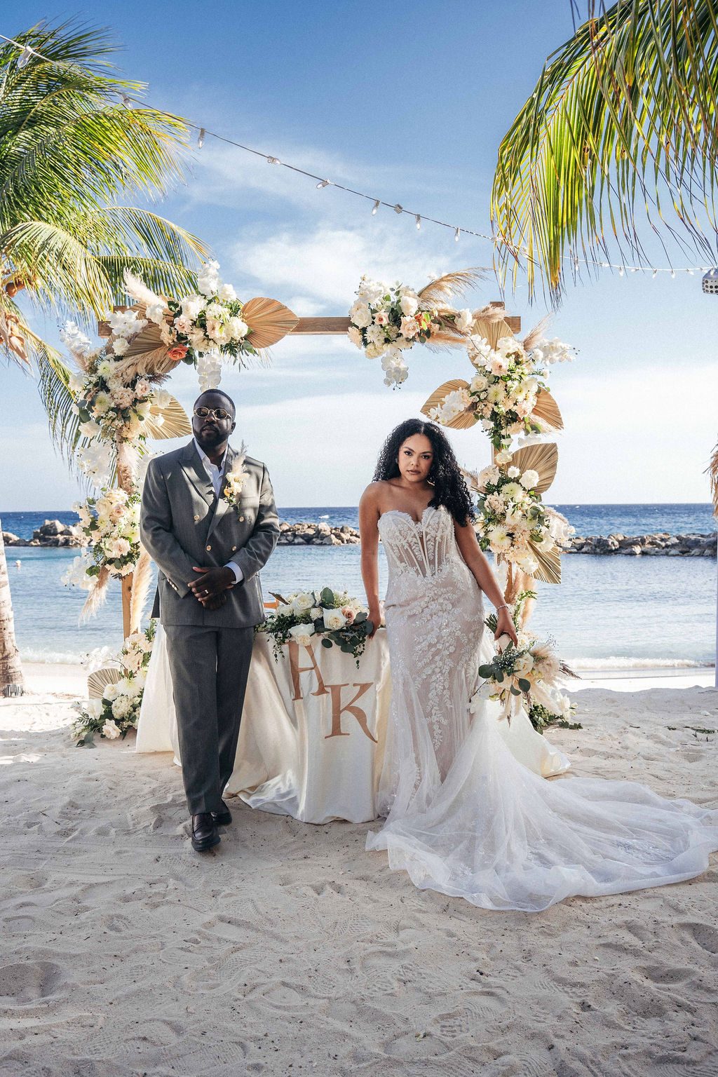 Kayla and Anthony stand in front of a floral arch at their wedding on the beach at Avila Beach Hotel, with the ocean in the background