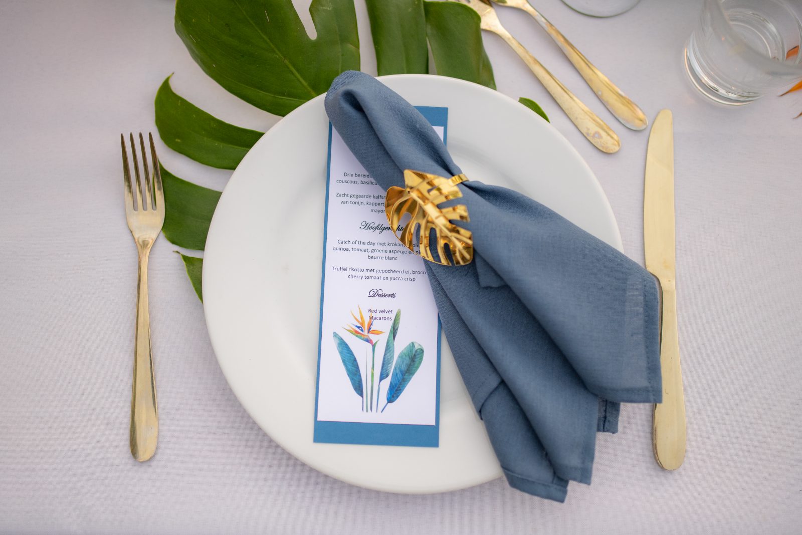 The table setting at Karakter, Curaçao features a plate with a blue napkin and menu, surrounded by gold utensils and tropical leaves