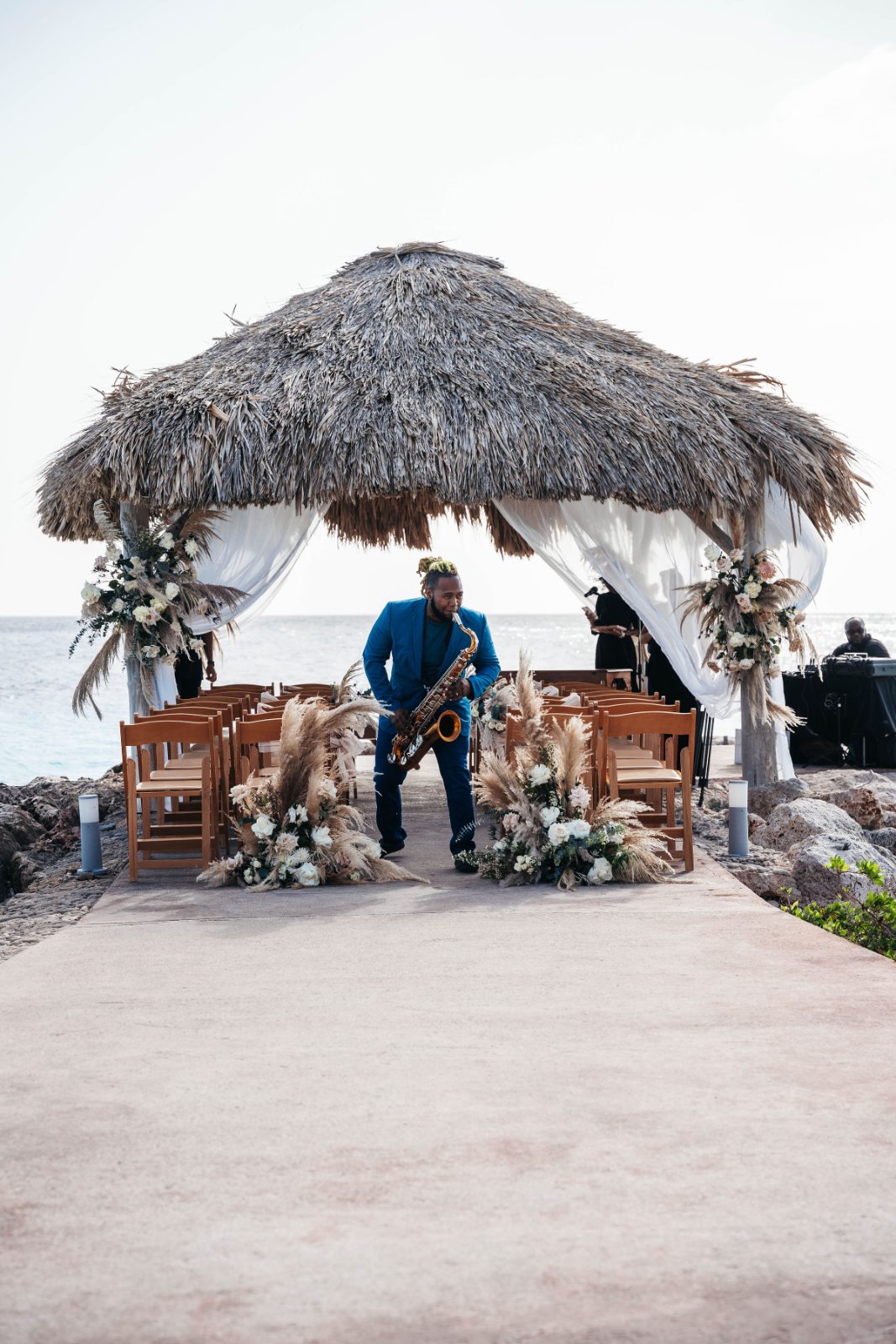 Kristal and Brittany's wedding ceremony is set at Karakter in Curaçao, featuring a saxophonist by a decorated thatched gazebo overlooking the ocean