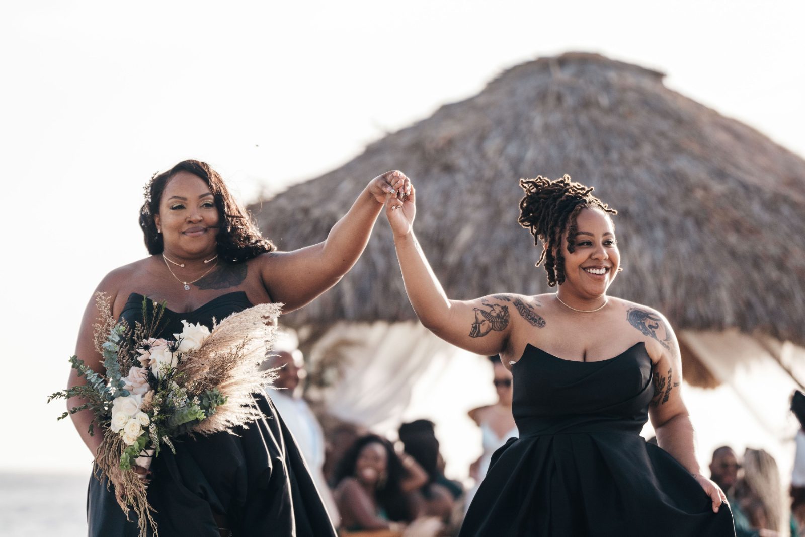 Kristal and Brittany walk down the beach at Karakter, Curaçao, with two bridesmaids holding hands, surrounded by guests under a thatched gazebo