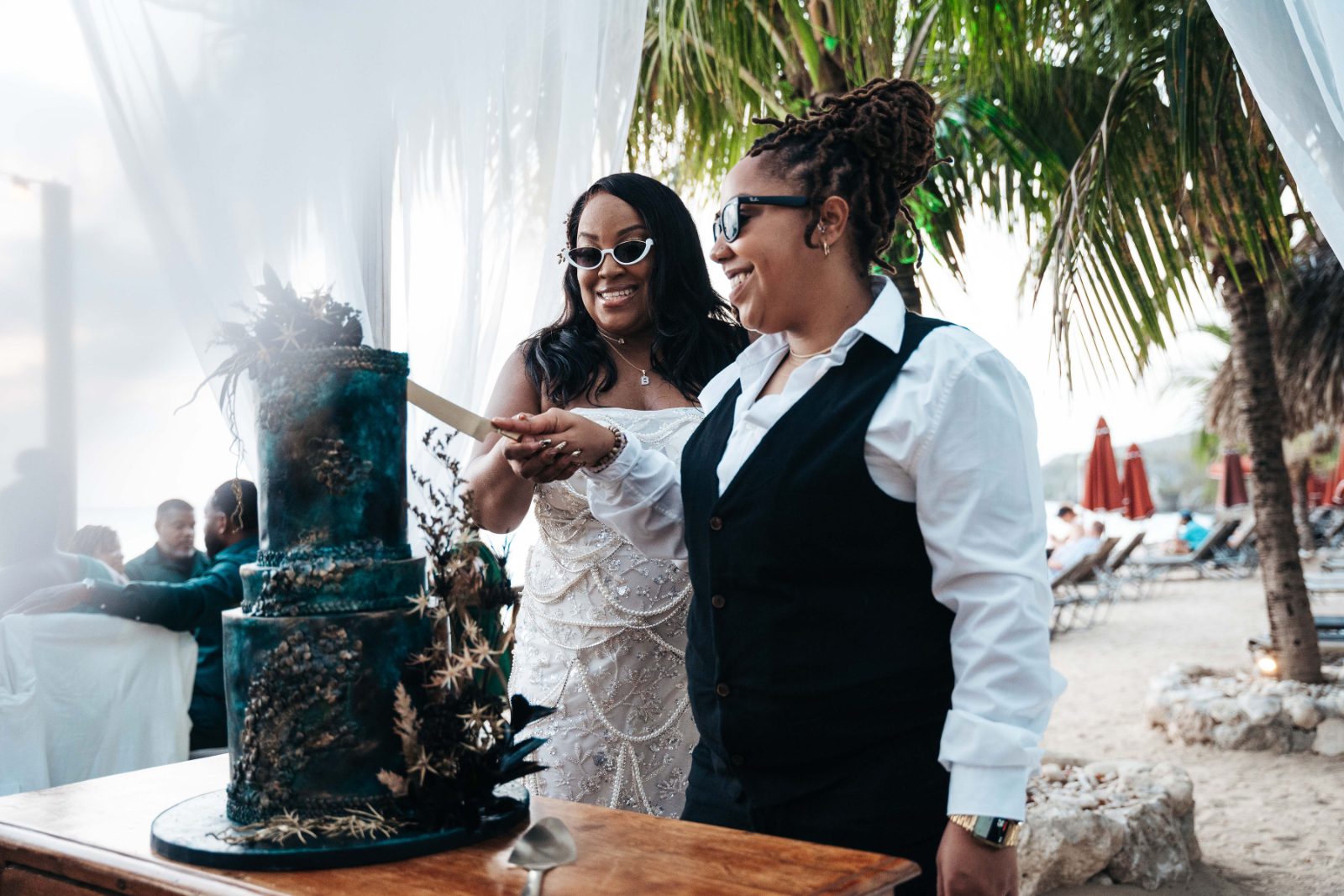 Kristal and Brittany are cutting a wedding cake at Karakter in Curaçao, surrounded by palm trees and guests in the background