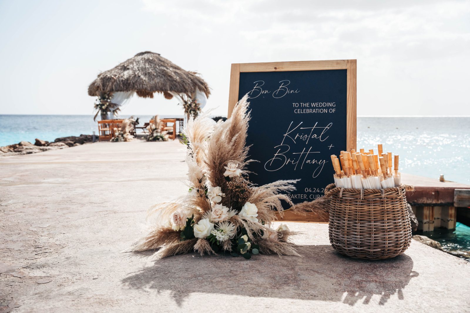 Kristal and Brittany's wedding setup at Karakter, Curaçao features a decorative sign and floral arrangements by the ocean
