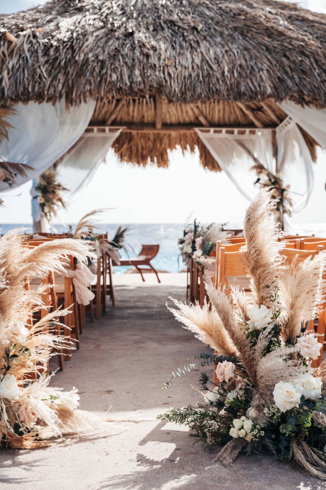 Kristal and Brittany are getting married under a thatched roof gazebo at Karakter, Curaçao, with floral decorations framing the aisle and seating