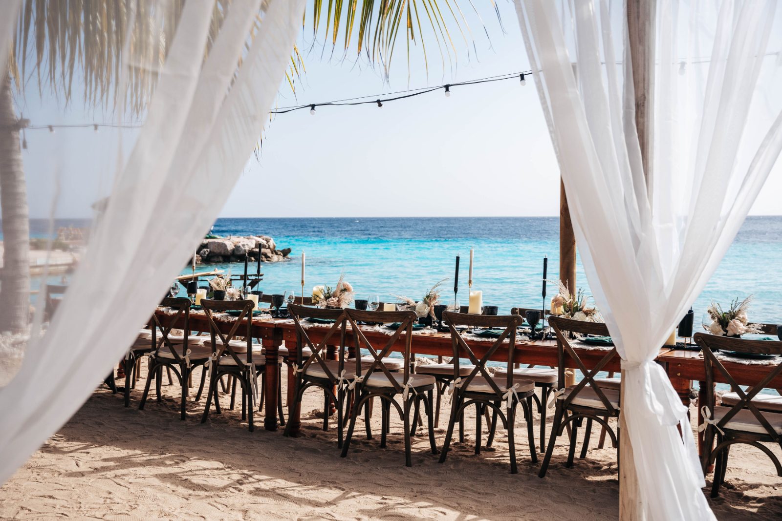 Kristal and Brittany's wedding reception is set at Karakter in Curaçao, featuring a long table with chairs beside a sandy beach and ocean view