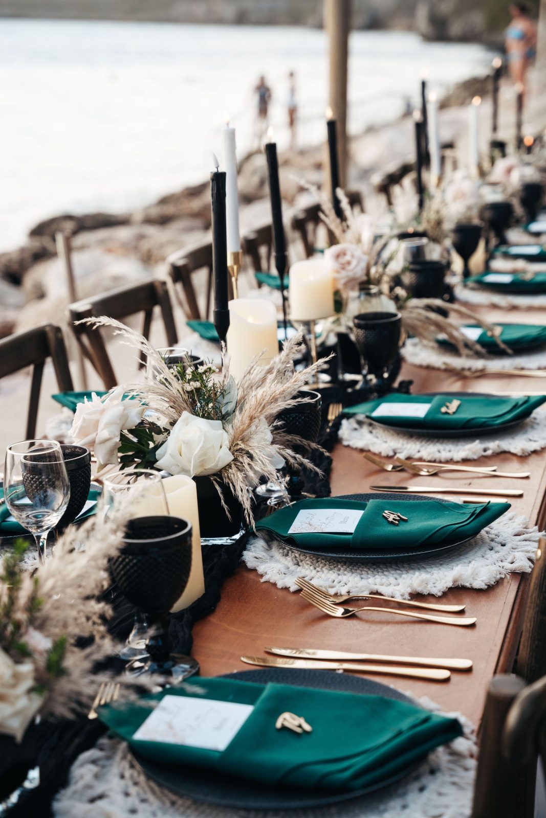 The wedding table features green tableware, black candles, and floral arrangements with a beachfront setting in Curaçao, while guests can be seen in the background
