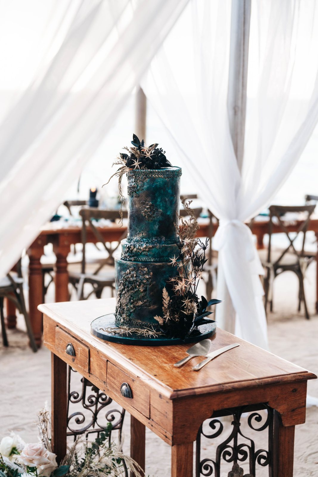Kristal and Brittany's wedding cake is displayed on a wooden table at Karakter in Curaçao, with decorative elements and a beach view in the background