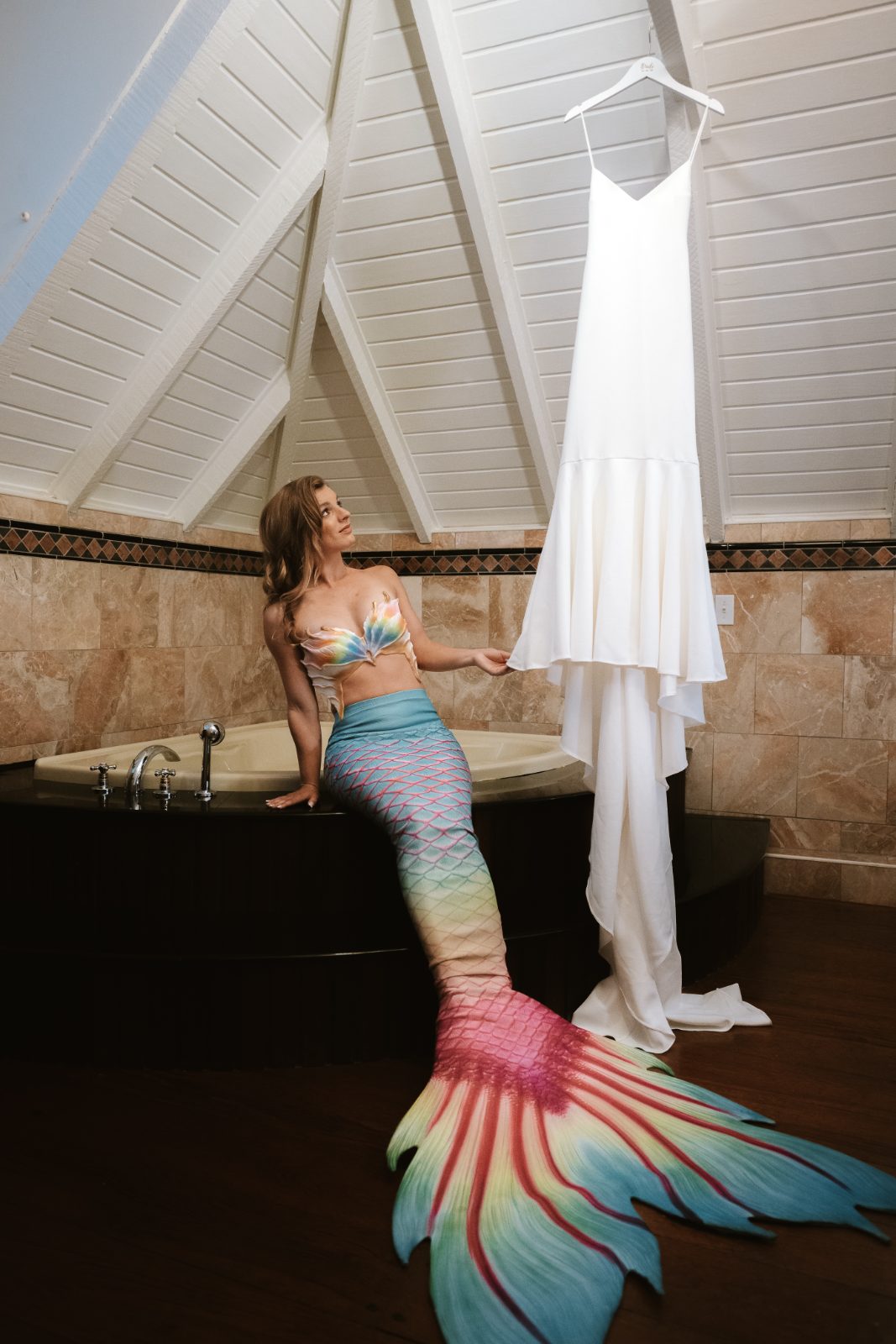 Megan, wearing a colorful mermaid outfit, sits by a bathtub in a Curaçao venue while admiring her white wedding dress hanging nearby