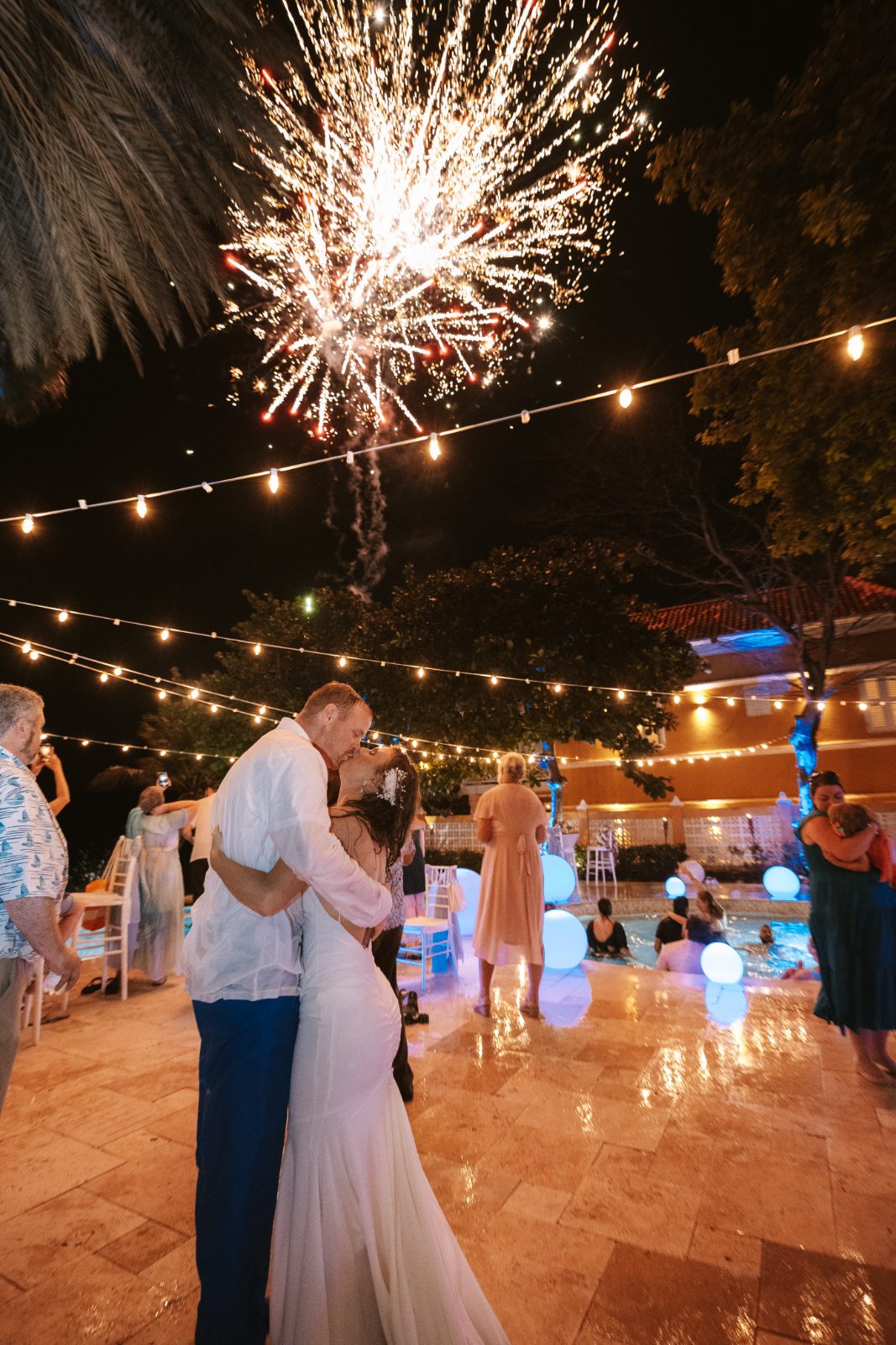 Megan and Nathan embrace on a lit outdoor patio in Curaçao as fireworks explode in the night sky above them