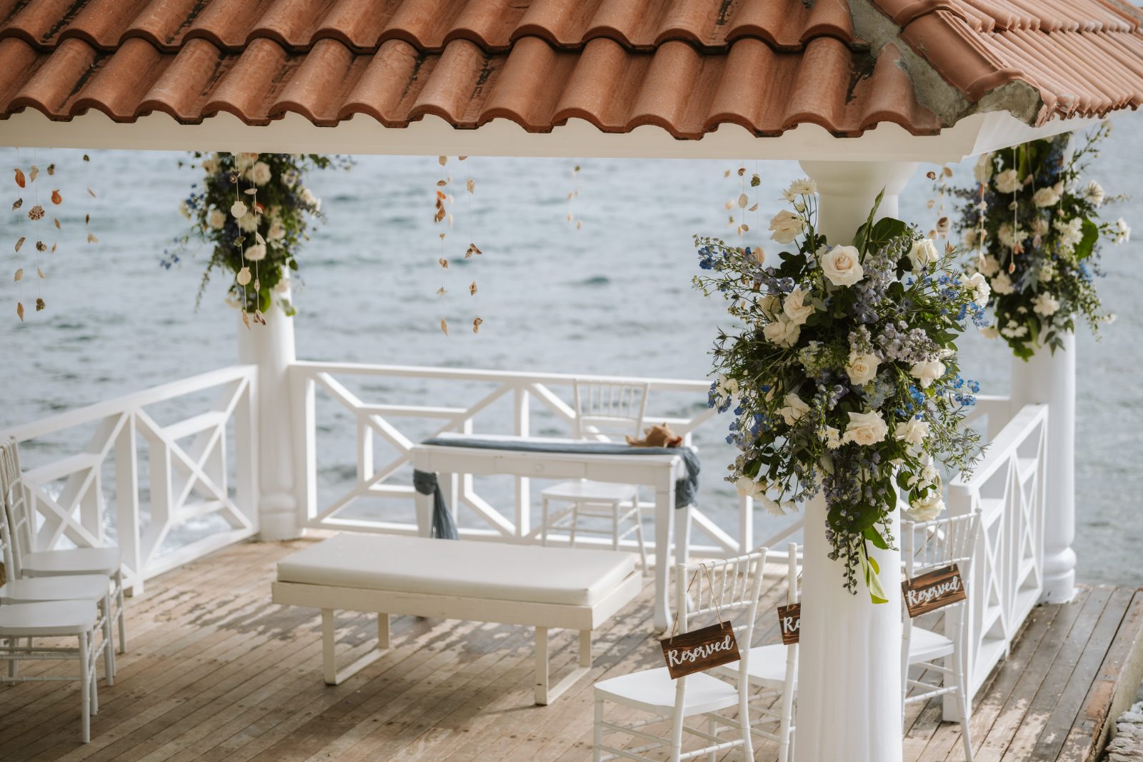 Megan and Nathan stand under a floral arch on a waterfront platform in Curaçao, surrounded by reserved chairs for their wedding ceremony
