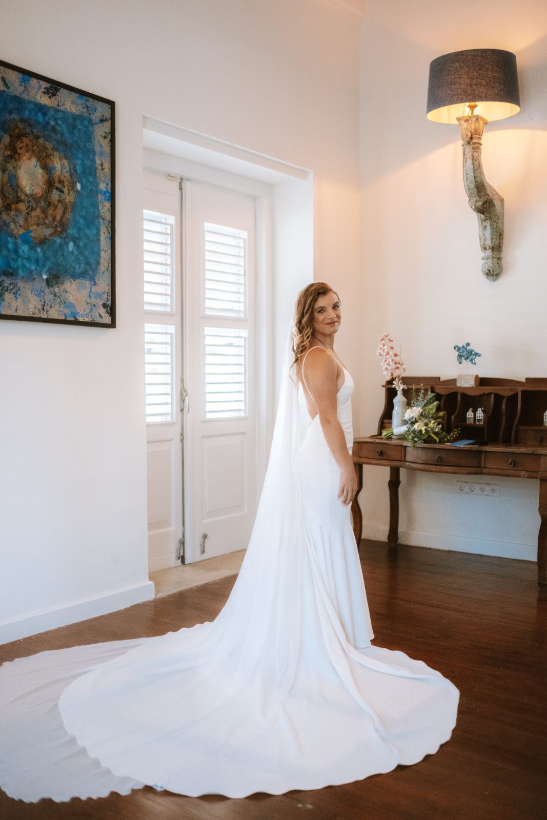 Megan stands in her wedding dress with a long train in a bright indoor space, preparing for her wedding to Nathan in Curaçao