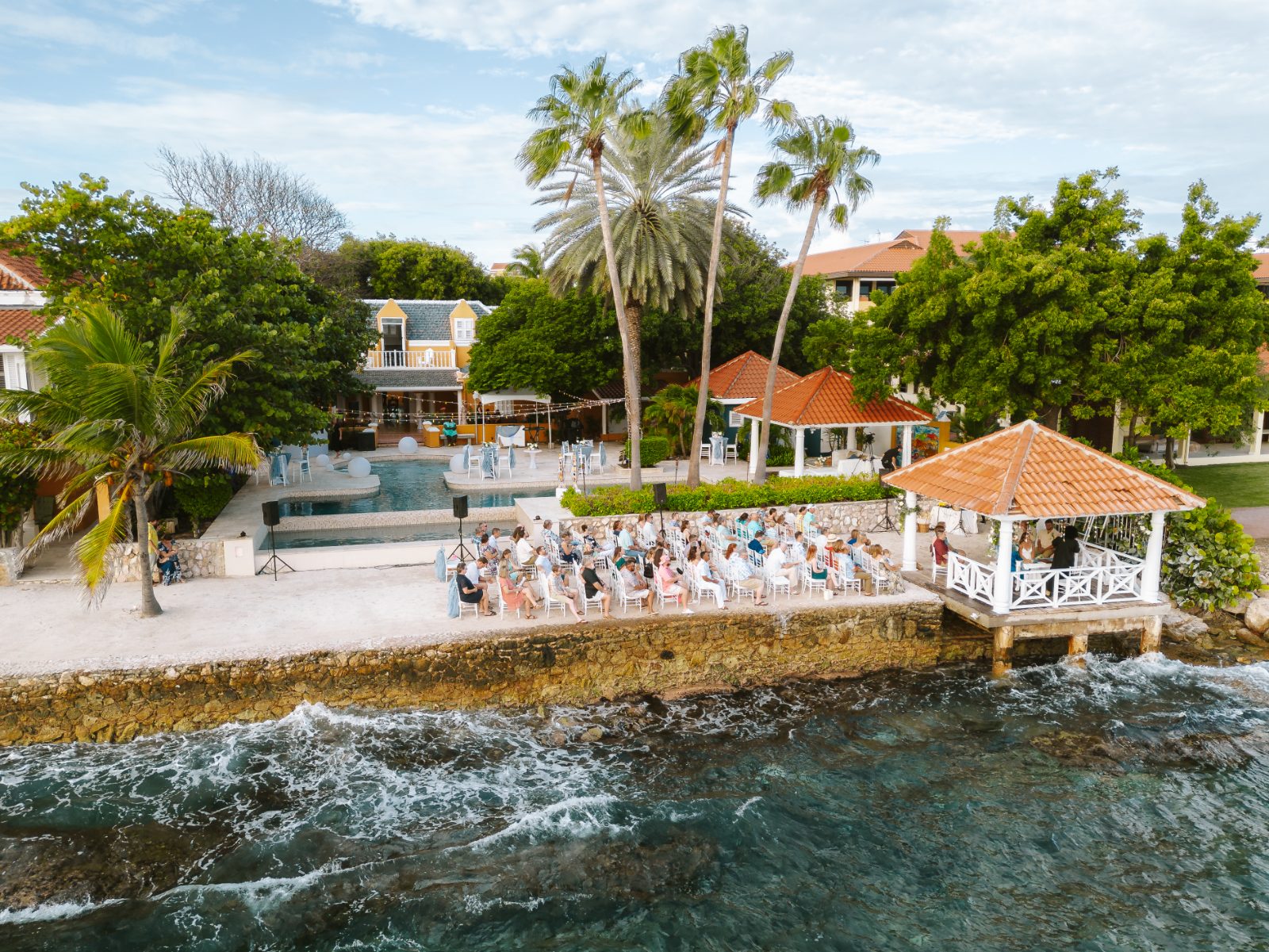Megan and Nathan are getting married outdoors by the water in Curaçao, surrounded by guests seated near a gazebo and tropical trees