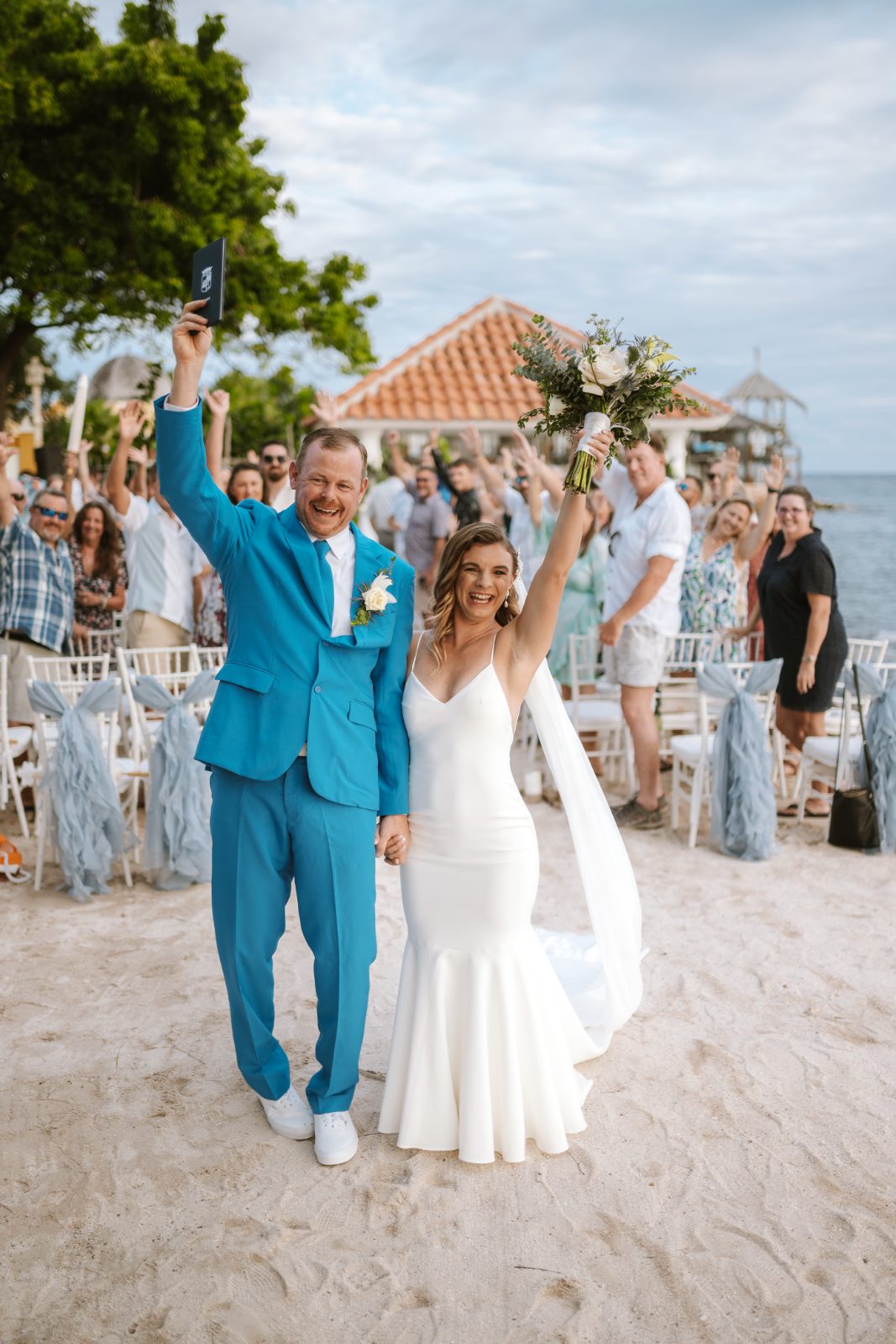 Megan and Nathan stand on the beach in Curaçao, celebrating their wedding with raised arms, while guests cheer in the background