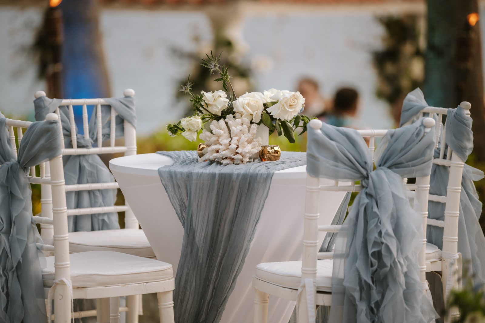 Megan and Nathan are at their wedding in Curaçao, with decorated chairs and a table featuring flowers and coral