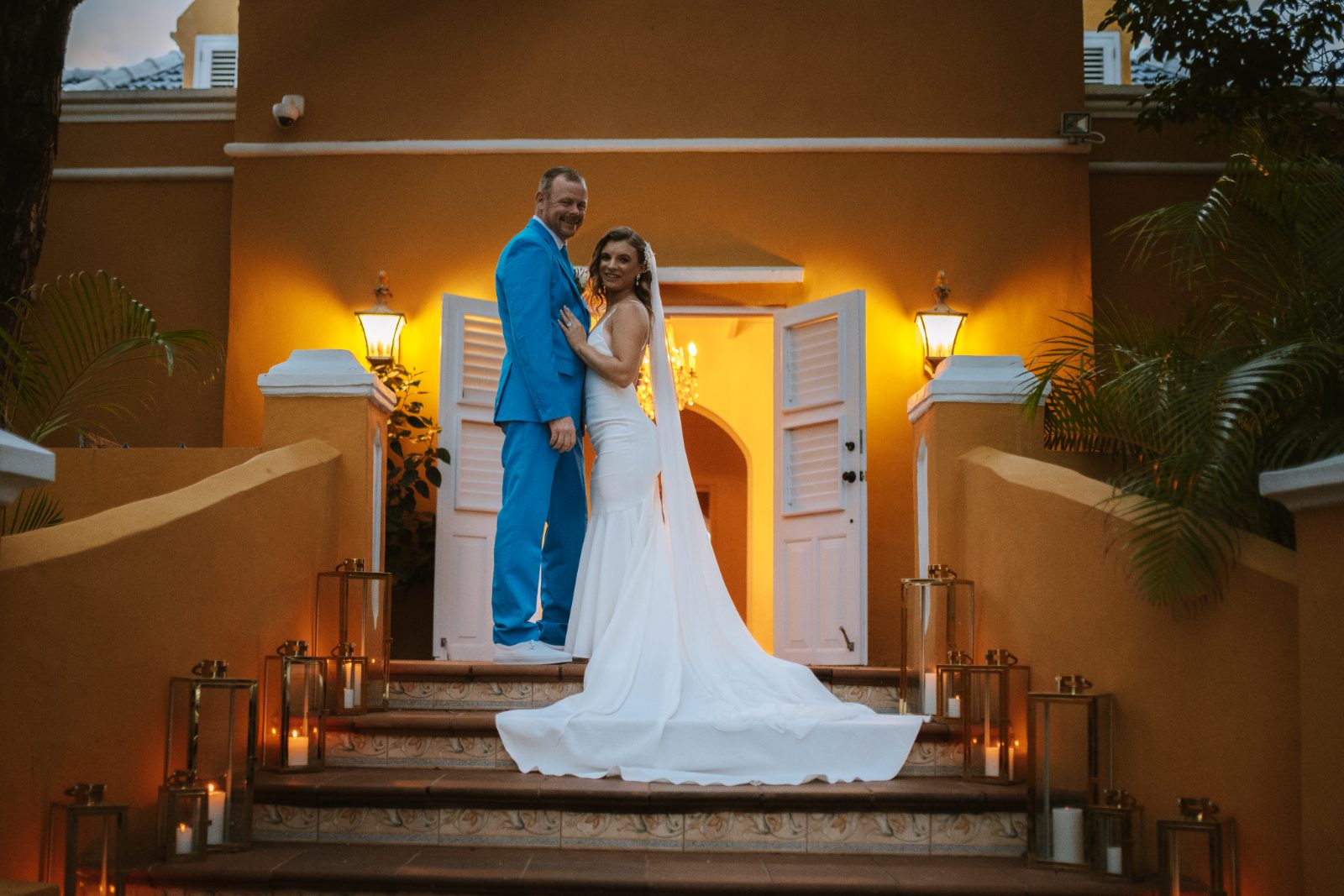 Megan and Nathan pose together on the steps of a yellow building in Curaçao, surrounded by lit lanterns and a warm evening ambiance