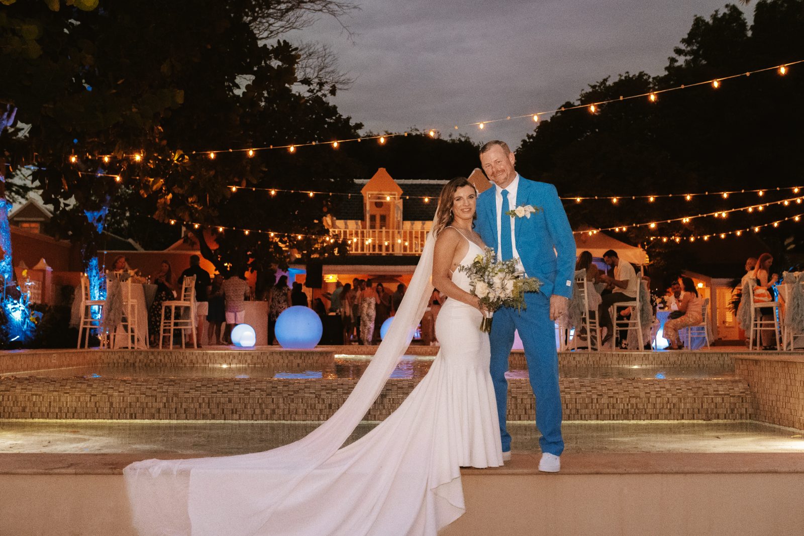 Megan and Nathan pose together by a fountain at their wedding in Curaçao, with guests and decorations visible in the background