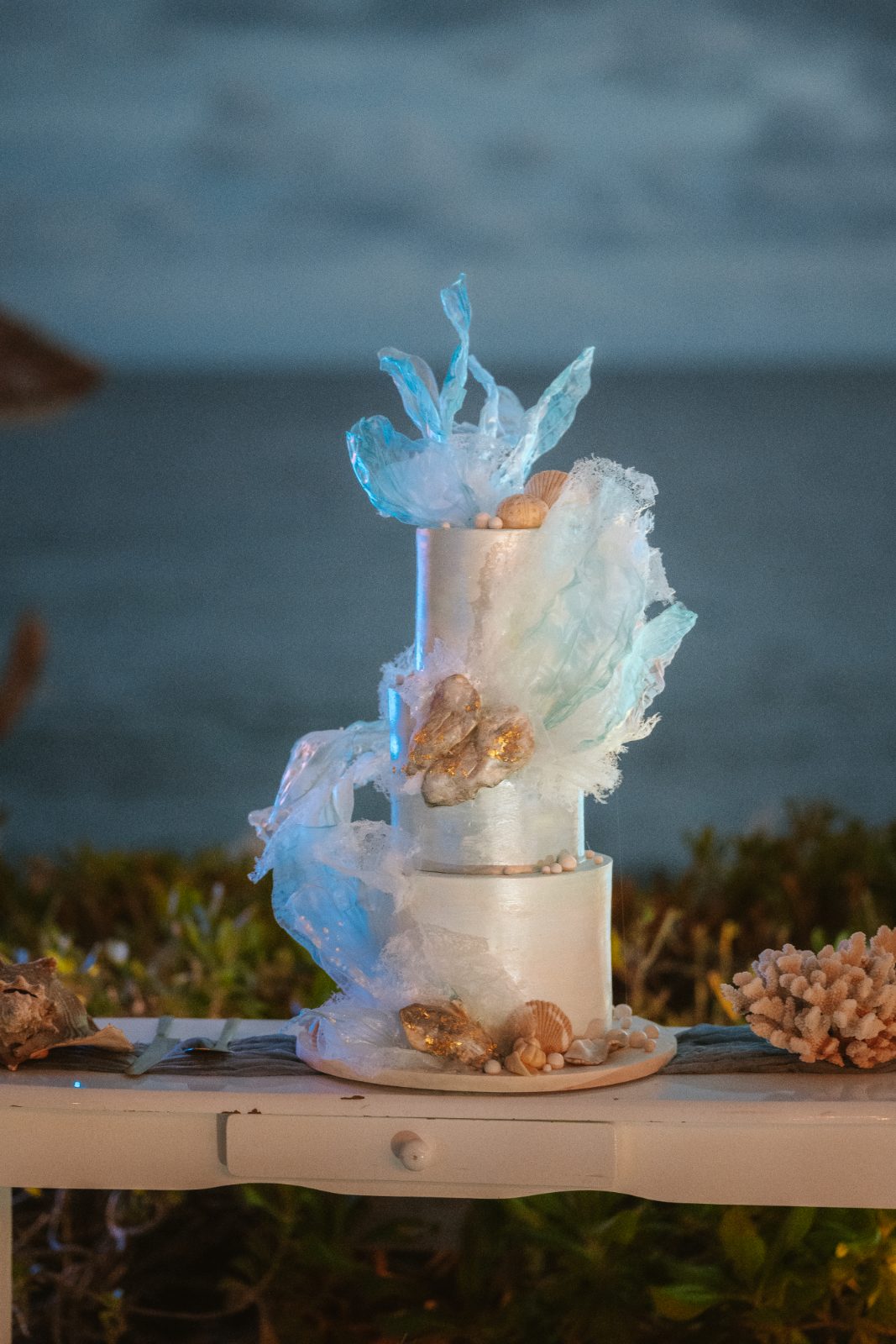 The wedding cake, decorated with shells and sea-themed elements, sits on a table overlooking the ocean in Curaçao, with a coastal backdrop