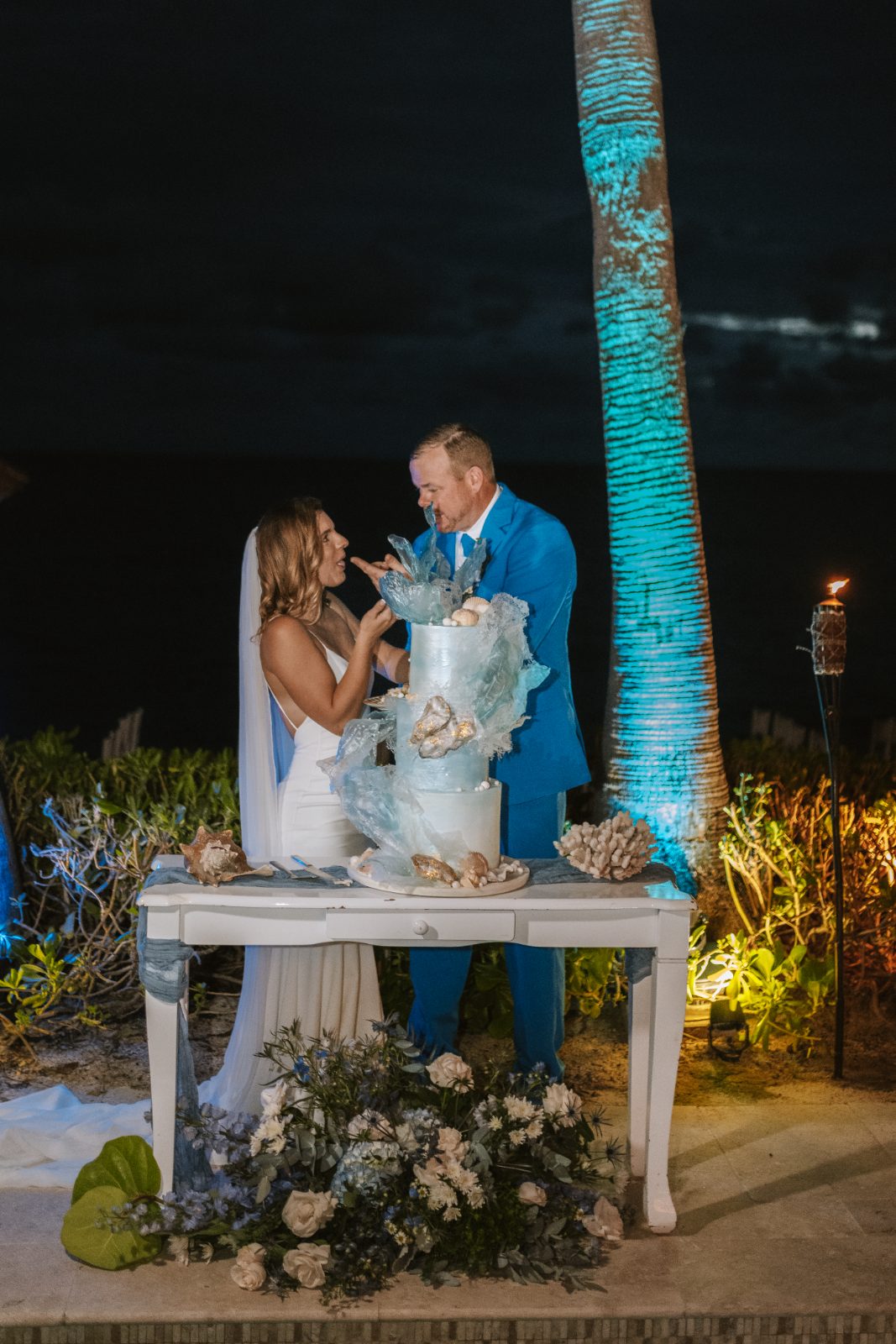 Megan and Nathan stand by their wedding cake at night in Curaçao, surrounded by tropical plants and illuminated by soft lighting