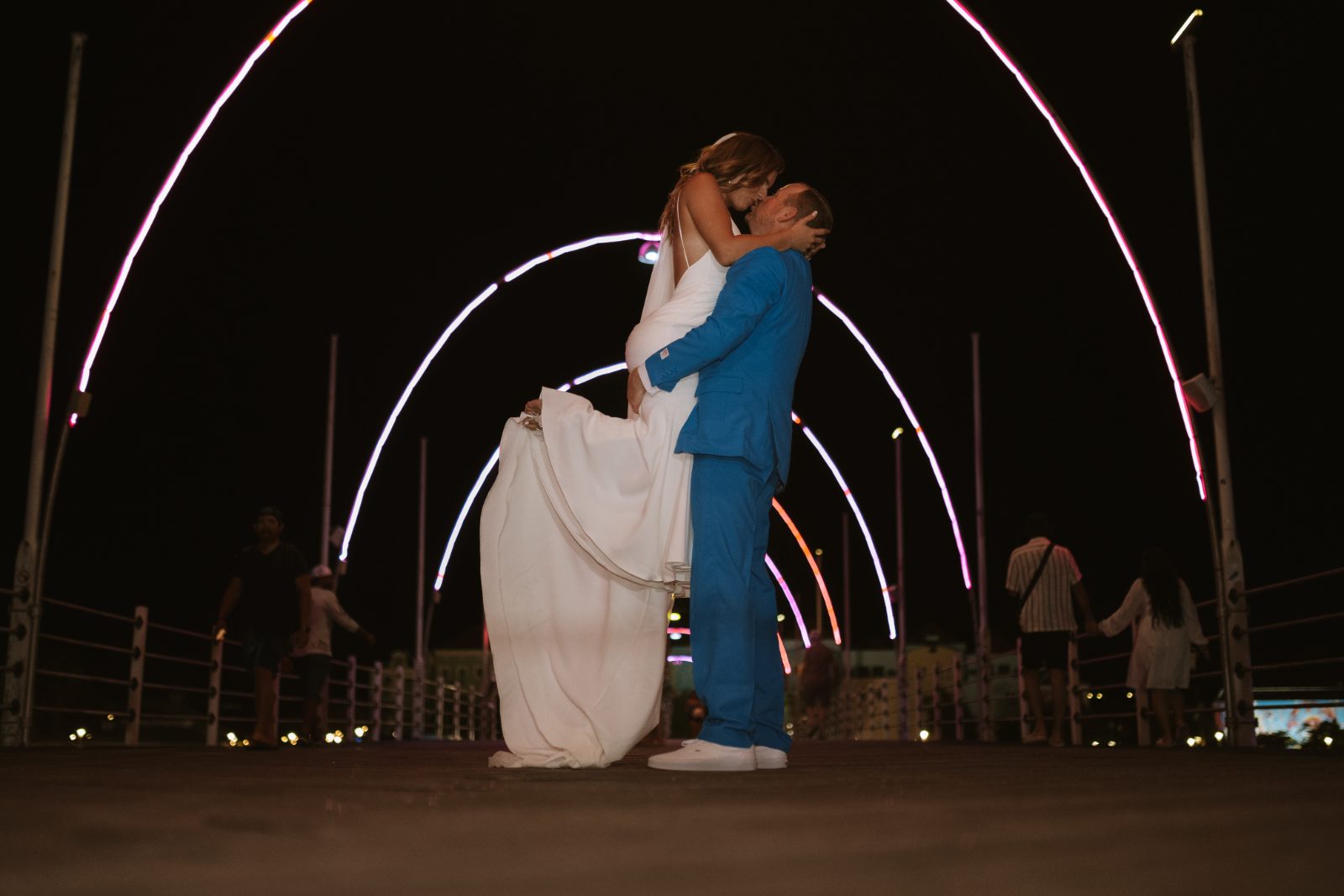 Megan and Nathan embrace on a brightly lit pier in Curaçao, with colorful lights framing the scene and guests visible in the background