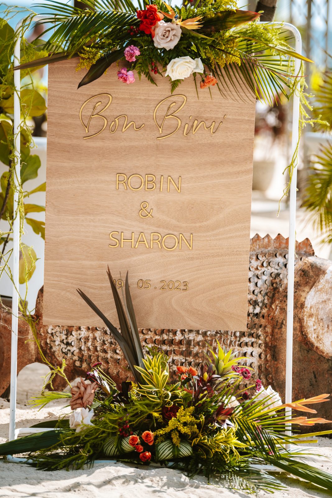 A wooden wedding sign at MOOD Beach, Curaçao, displays the names Robin and Sharon, surrounded by flowers and tropical foliage