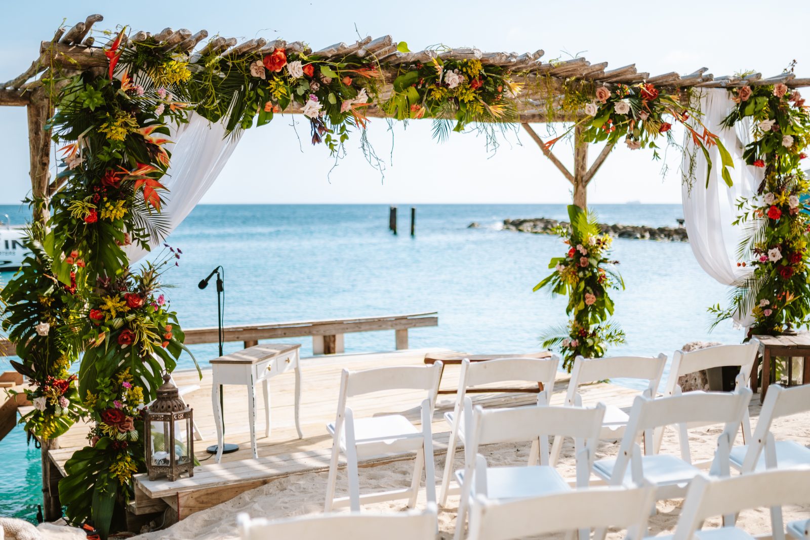 Sharon and Robin's wedding ceremony is set at MOOD Beach in Curaçao, featuring a decorated arch by the water with white chairs arranged for guests