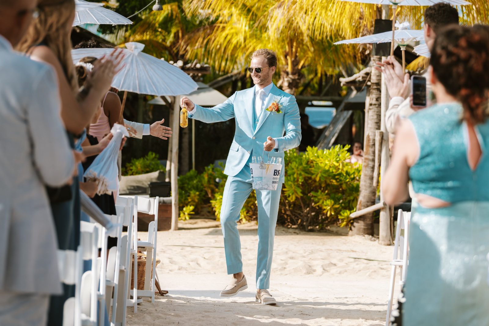 Sharon and Robin's wedding at MOOD Beach, Curaçao, features a groom in a light blue suit walking down the sandy aisle, greeted by guests