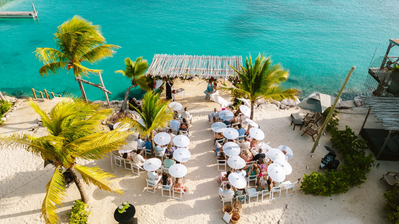 Sharon and Robin are getting married at MOOD Beach in Curaçao, surrounded by guests seated under umbrellas by the ocean