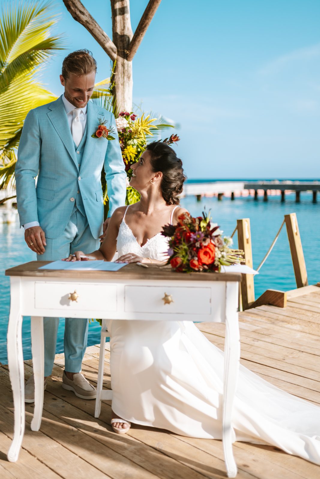 Sharon and Robin are standing at a table by the waterfront at MOOD Beach, Curaçao, during their wedding ceremony