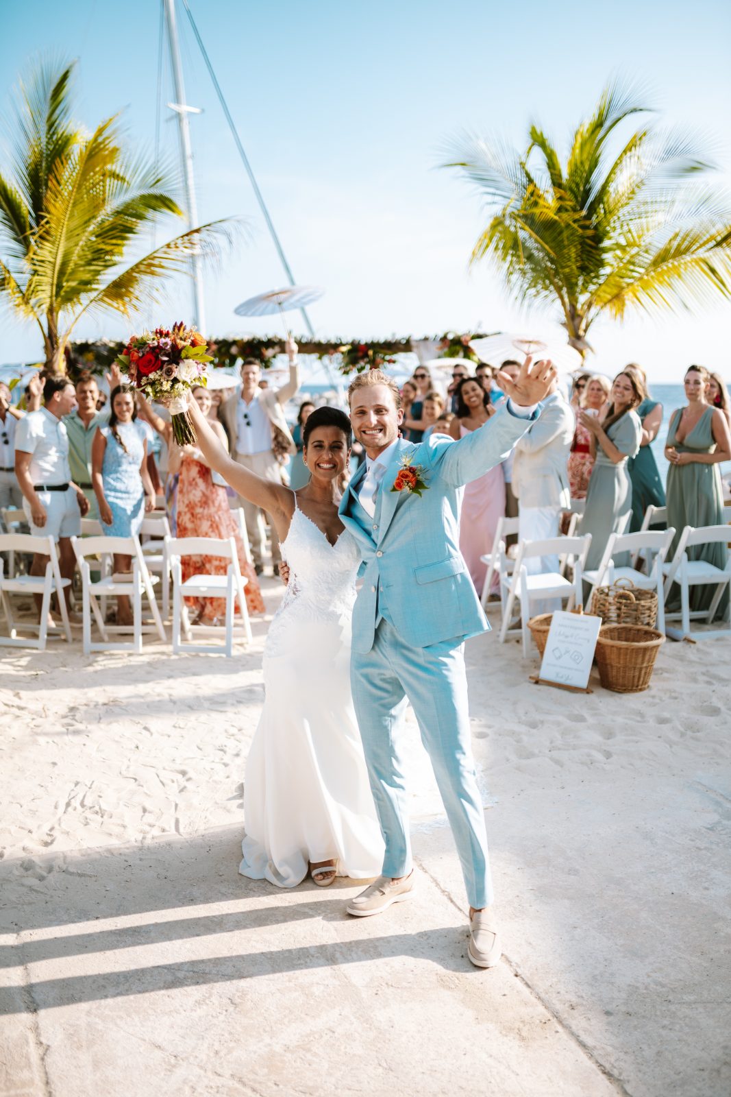 Sharon and Robin celebrate their wedding at MOOD Beach in Curaçao, surrounded by guests and palm trees, with Robin waving and Sharon holding a bouquet