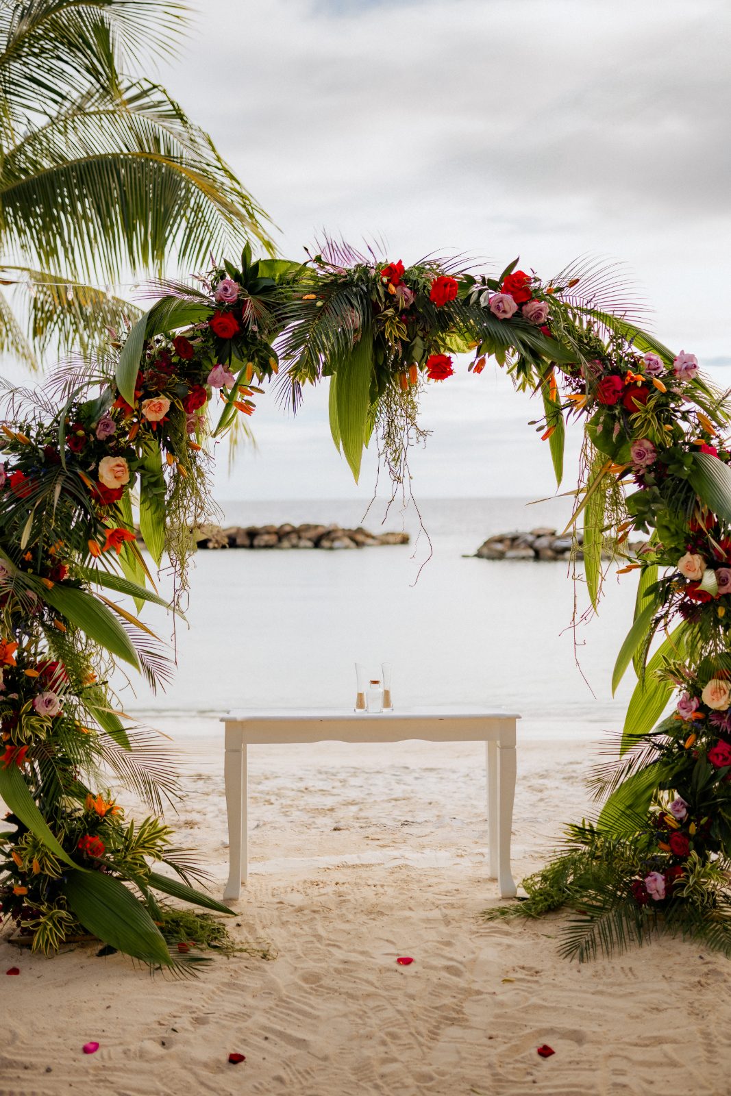Stephanie and Simon are standing under a floral arch by the beach at Avila Beach Hotel, with the ocean visible in the background