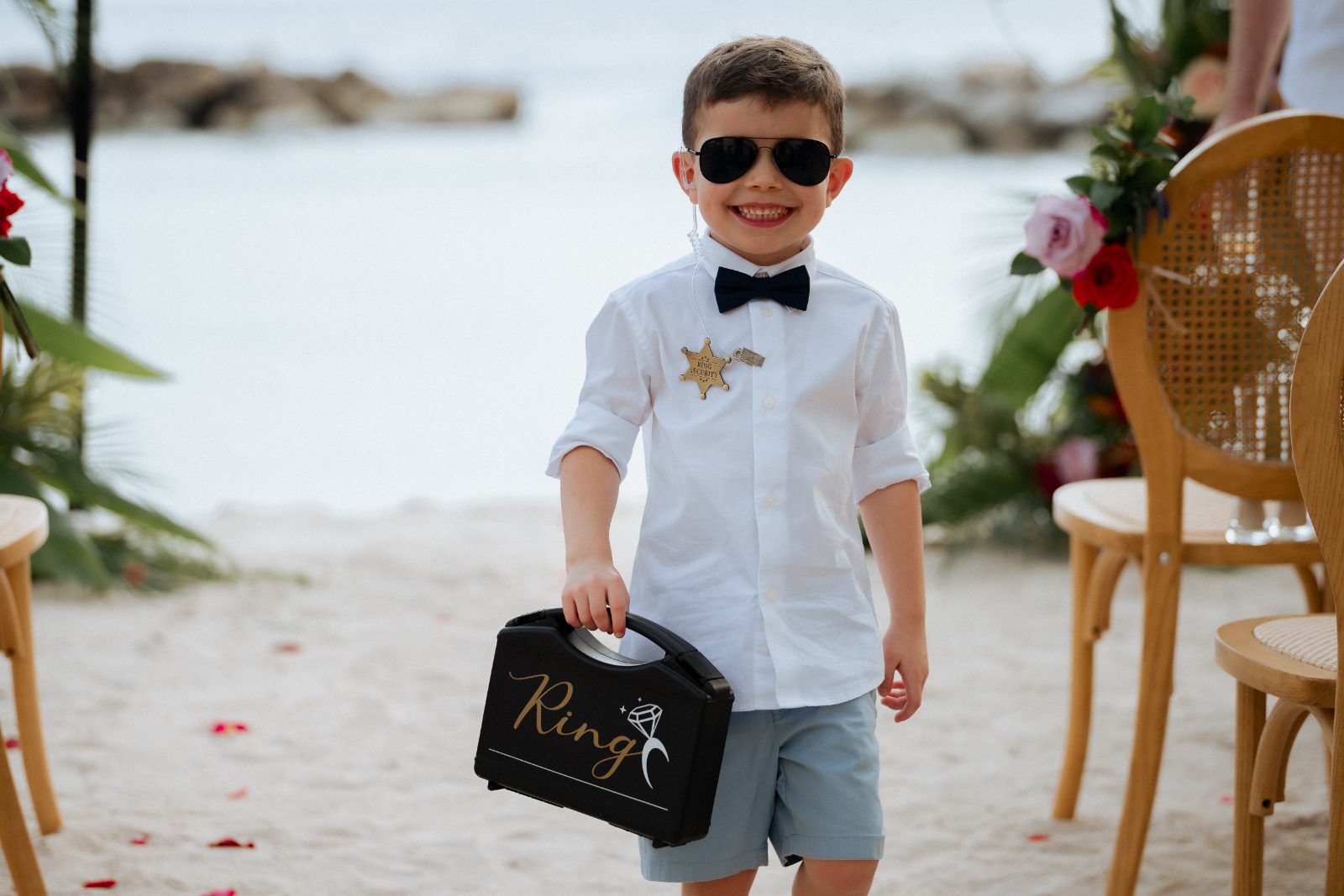 A young boy in sunglasses and a white shirt carries a black box labeled "Ring" at a beach wedding in Curaçao