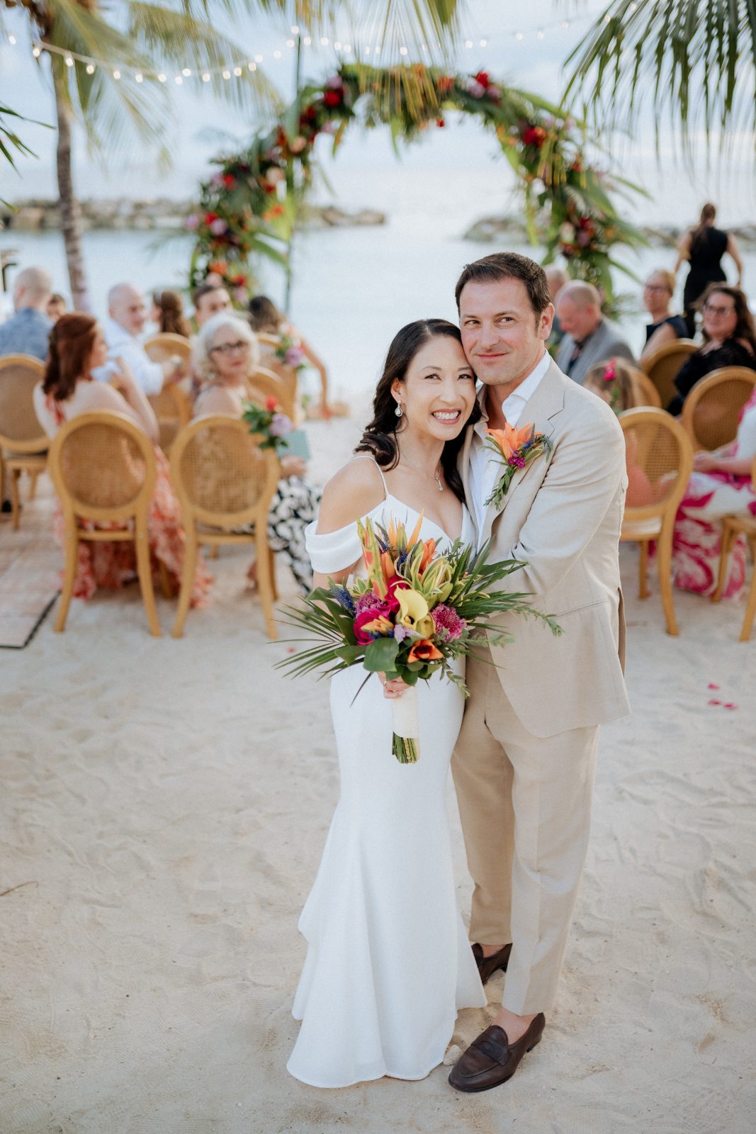 Stephanie and Simon stand together after their wedding ceremony at Avila Beach Hotel in Curaçao, with guests seated in the background and tropical decorations around them