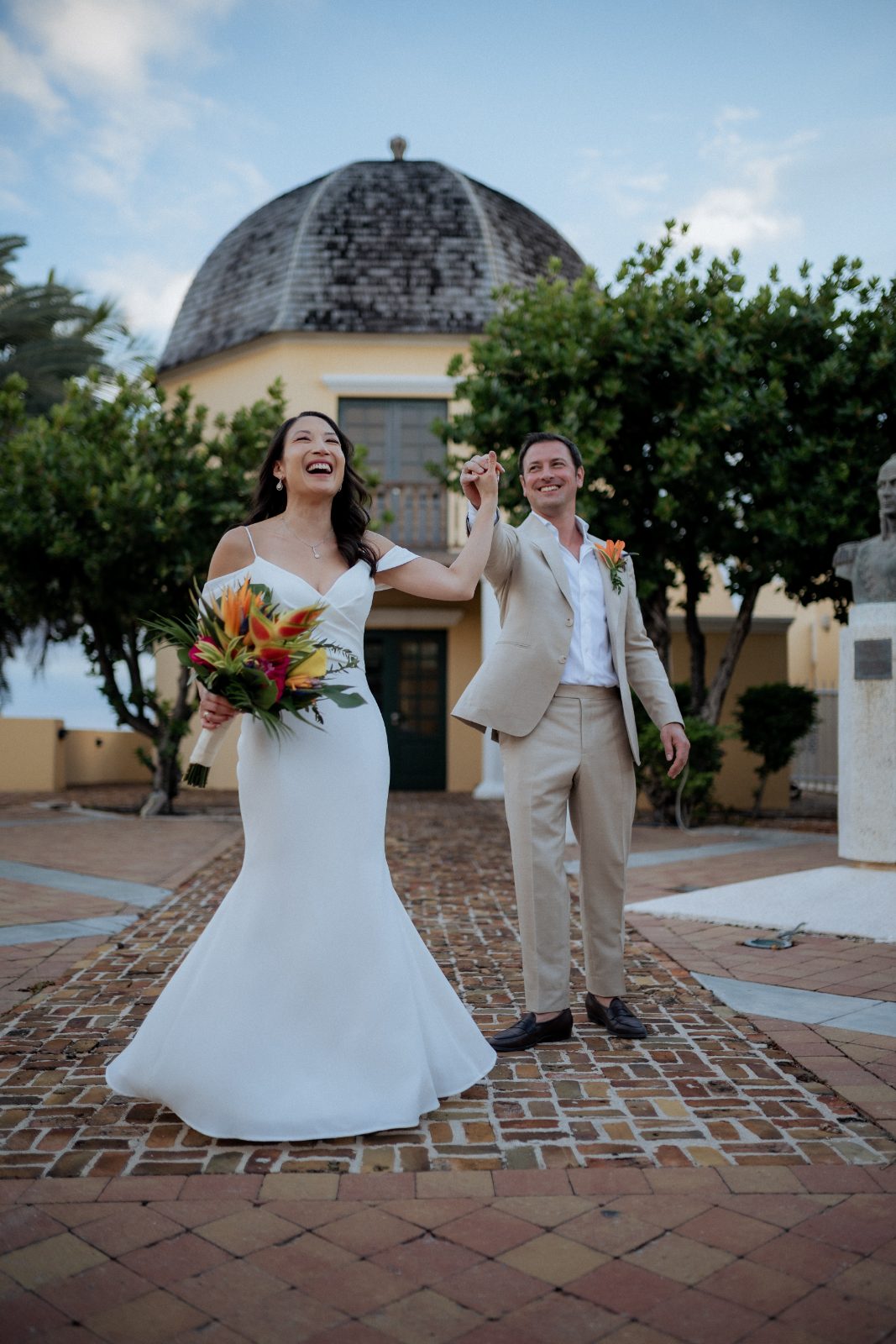 Stephanie and Simon celebrate their wedding at Avila Beach Hotel in Curaçao, standing on a brick path beside greenery, holding hands and smiling
