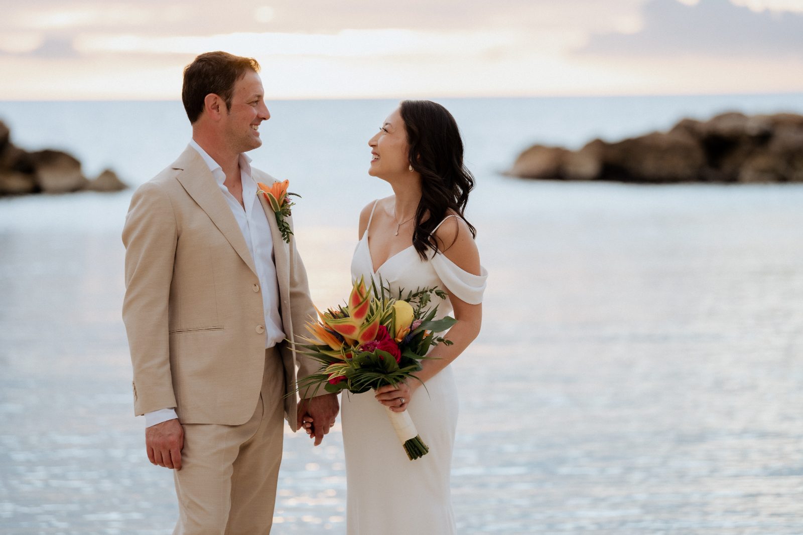 Stephanie and Simon hold hands on a beach at Avila Beach Hotel in Curaçao, with a bouquet of tropical flowers in Stephanie's hand and water behind them