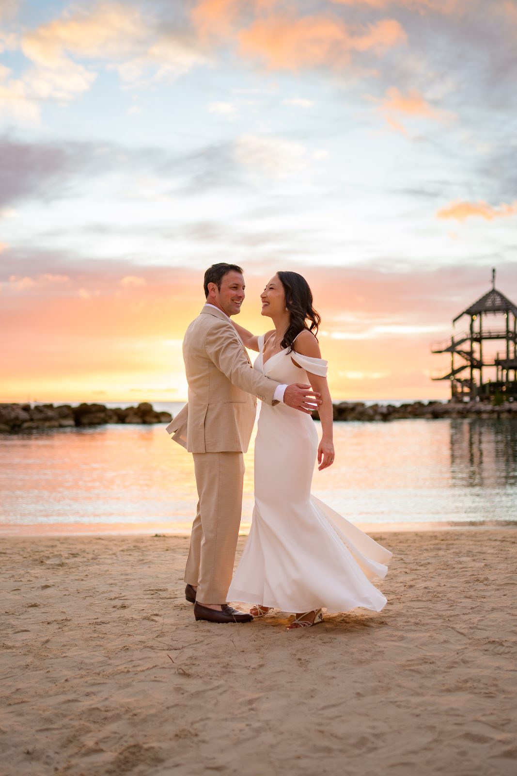 Stephanie and Simon stand together on the beach at Avila Beach Hotel in Curaçao, with a sunset and water in the background