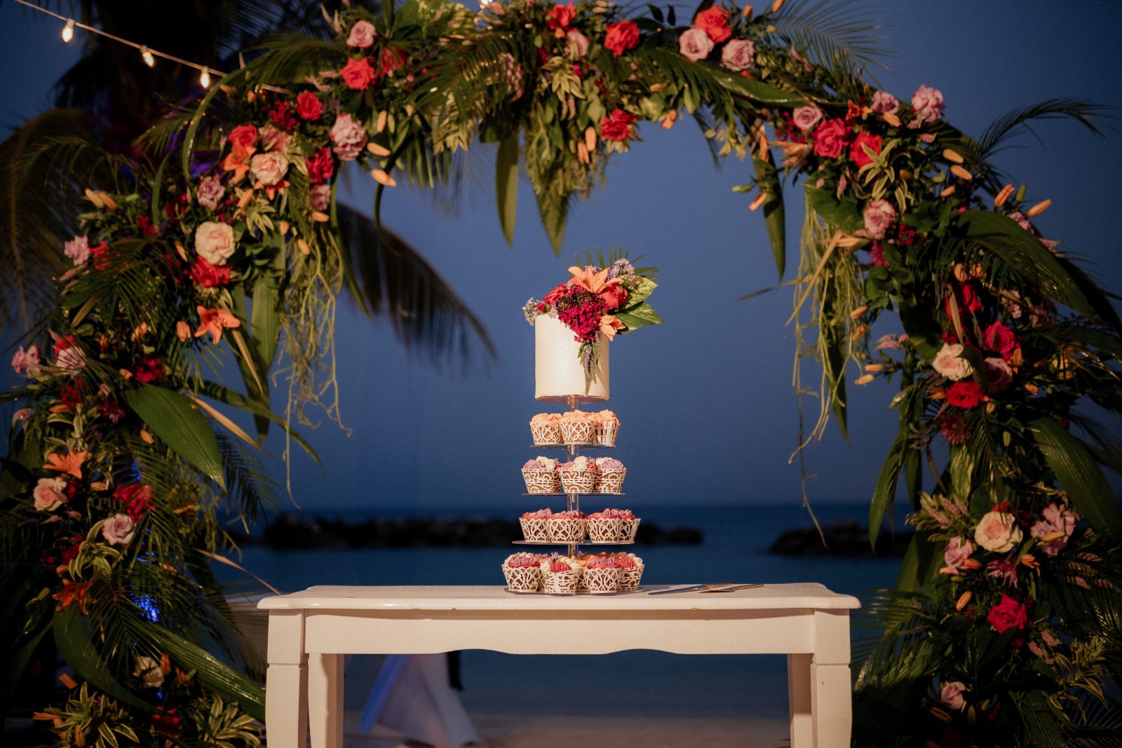 Stephanie and Simon's wedding features a tiered cupcake display set in front of a floral arch at Avila Beach Hotel, Curaçao, by the ocean